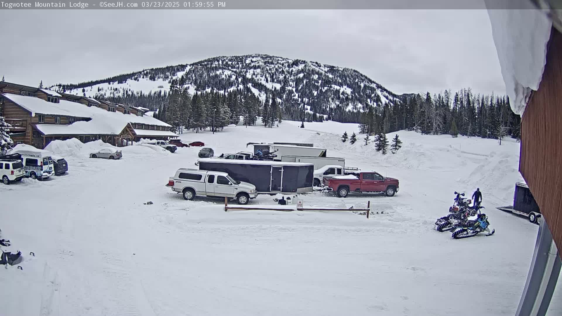 A snow-covered parking lot with several vehicles, including trucks and trailers, is situated in front of a lodge, with a snow-covered mountain in the background under an overcast sky.