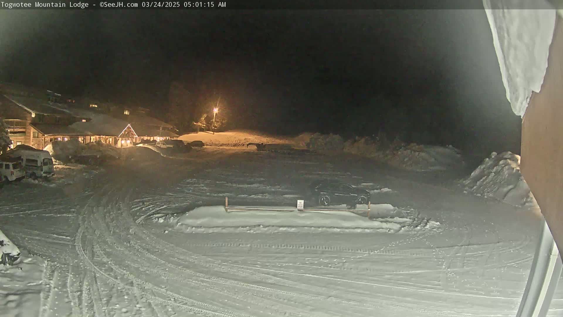A snow-covered parking lot at night, illuminated by a single lamp, with a lodge visible in the background under a dark sky.