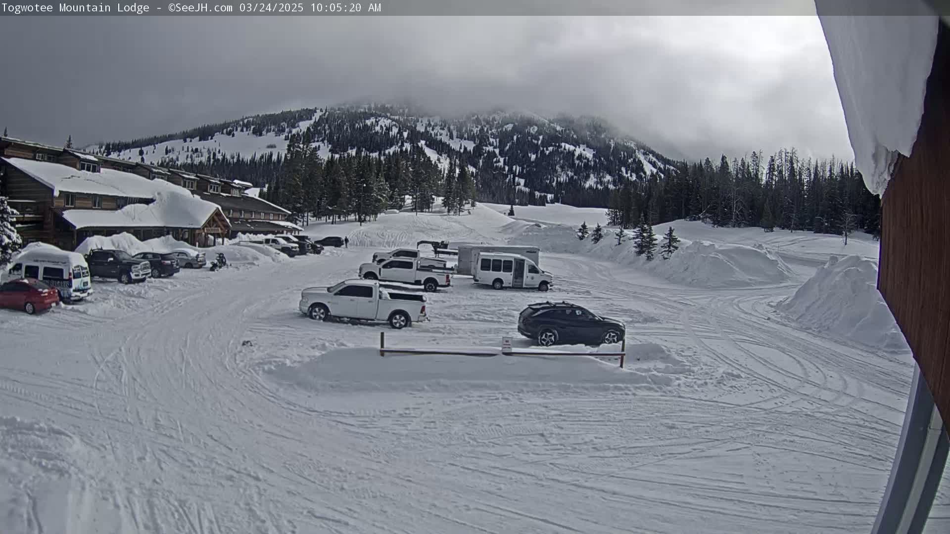 A snow-covered parking lot with several vehicles, including trucks and SUVs, is situated in front of a lodge nestled in a mountainous, snow-covered landscape under an overcast sky.