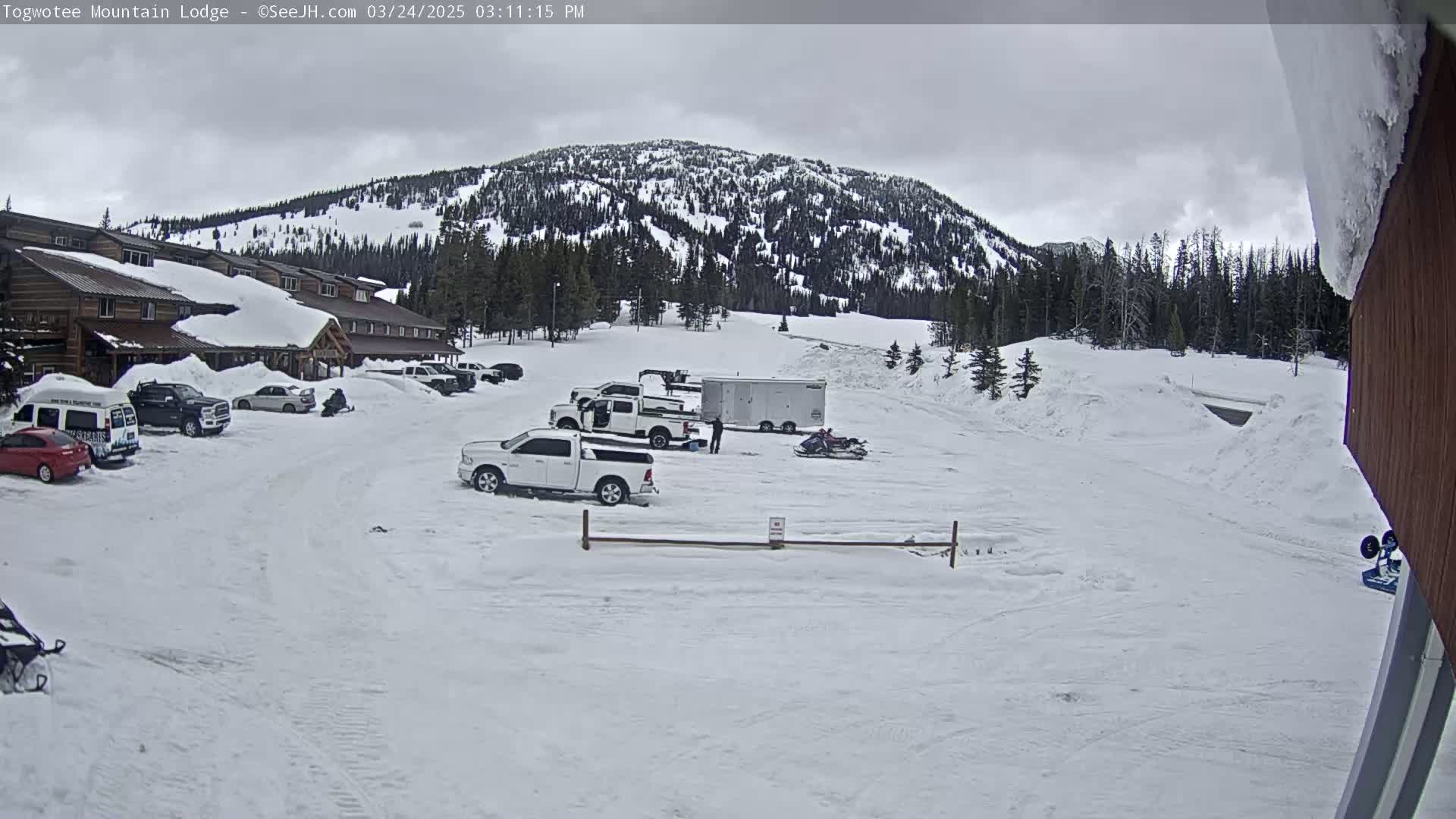 A snow-covered parking lot with several vehicles, including trucks and a van, sits in front of a snow-laden lodge, with a snow-covered mountain in the background under an overcast sky.