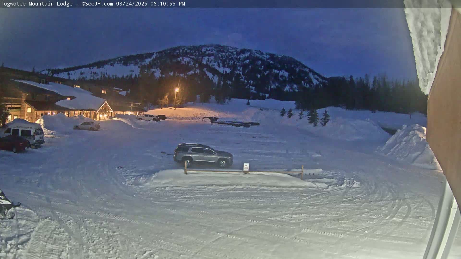 A snow-covered parking lot in front of a lodge sits beneath a twilight sky, with a mountain in the background.