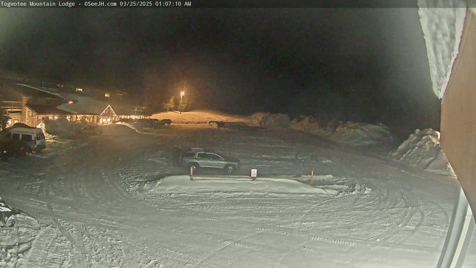 A snow-covered parking lot at night, illuminated by a single light, with a lodge and several vehicles visible.