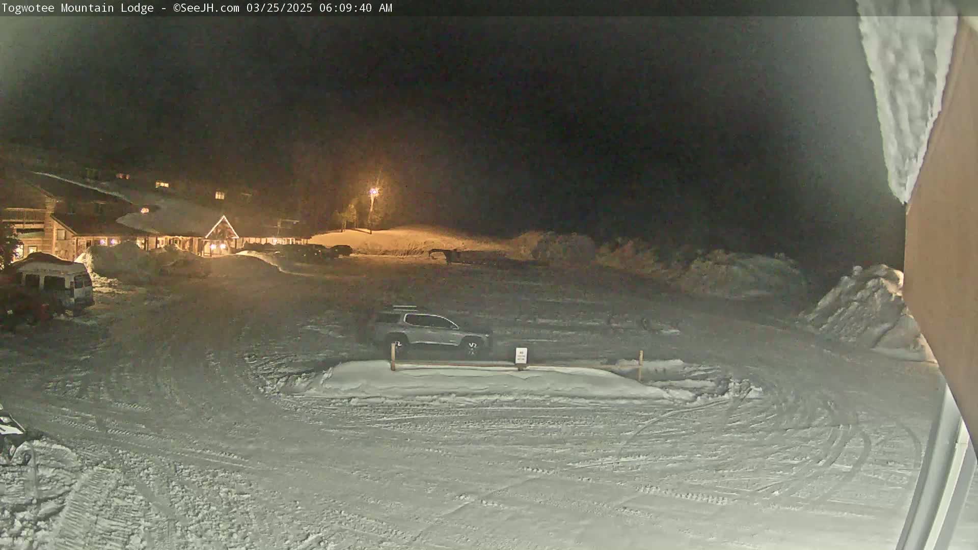 A snow-covered parking lot at night, illuminated by a single lamppost, shows a lodge in the background with several cars parked, and tire tracks in the snow.