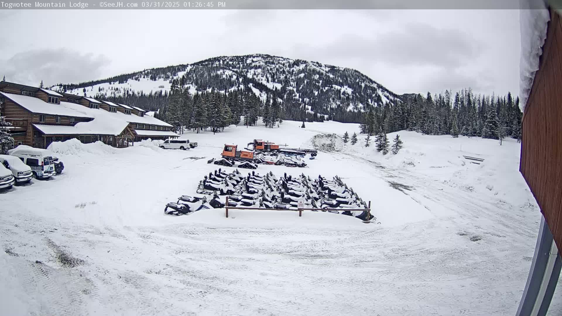 A snow-covered parking lot with numerous snowmobiles parked in front of a lodge, nestled at the base of a snow-dusted mountain range under an overcast sky.