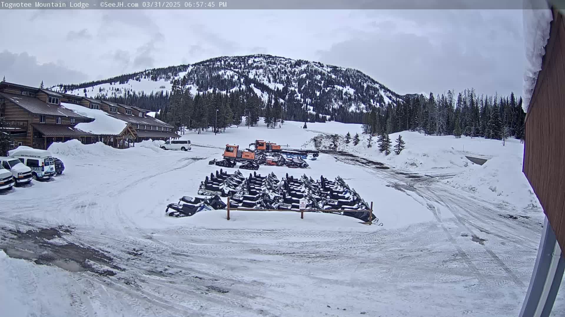 A snow-covered parking lot with numerous snowmobiles parked in front of a lodge and a snow-covered mountain in the background under an overcast sky.
