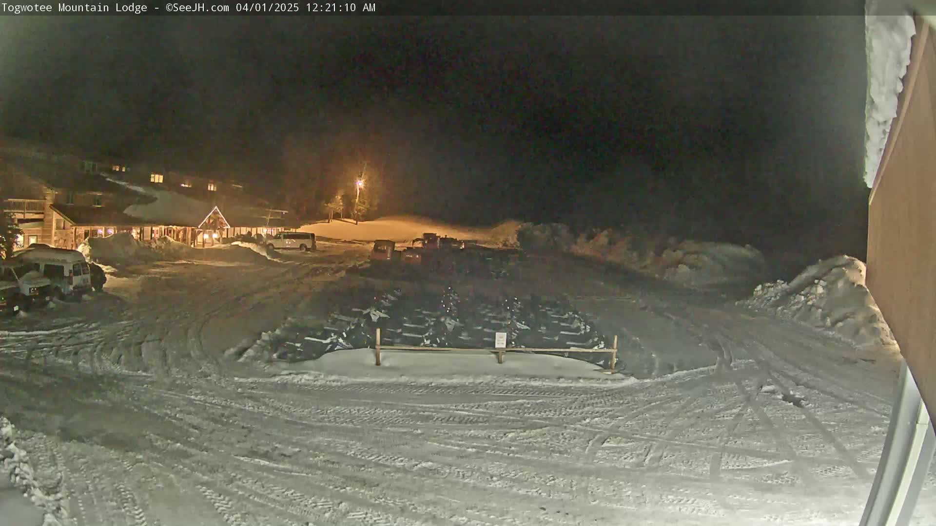 A snow-covered parking lot at night, with a lodge in the background, contains numerous snowmobiles parked behind a fence.