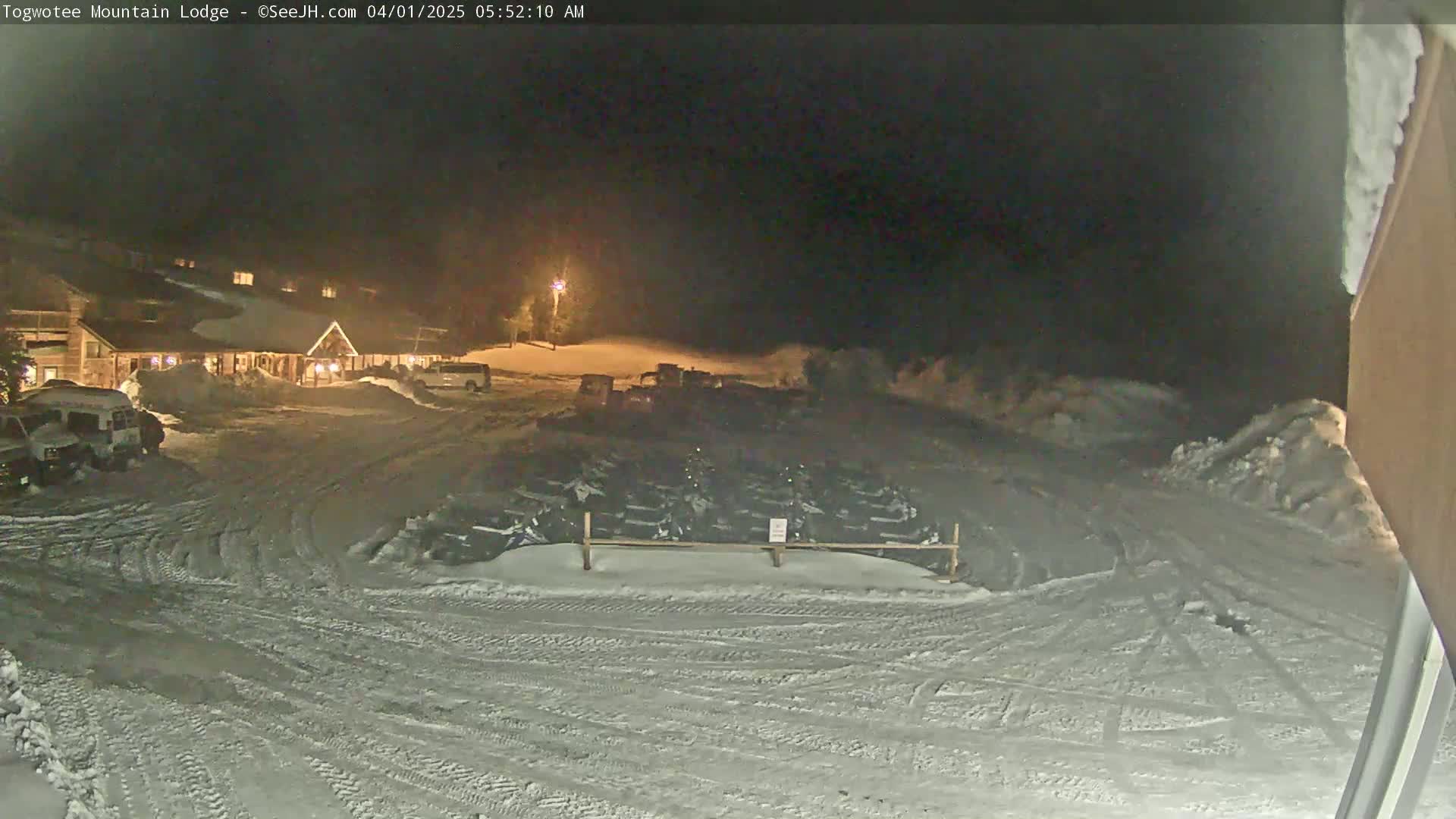 A snow-covered parking lot at night, with a lodge and numerous snowmobiles parked nearby under artificial light.