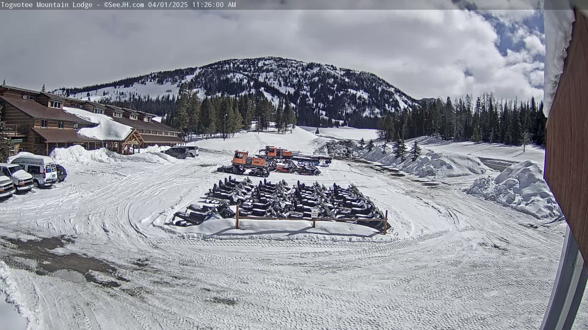 A snow-covered lodge parking lot with numerous snowmobiles parked, snowcats nearby, and a mountainous backdrop under a partly cloudy sky.