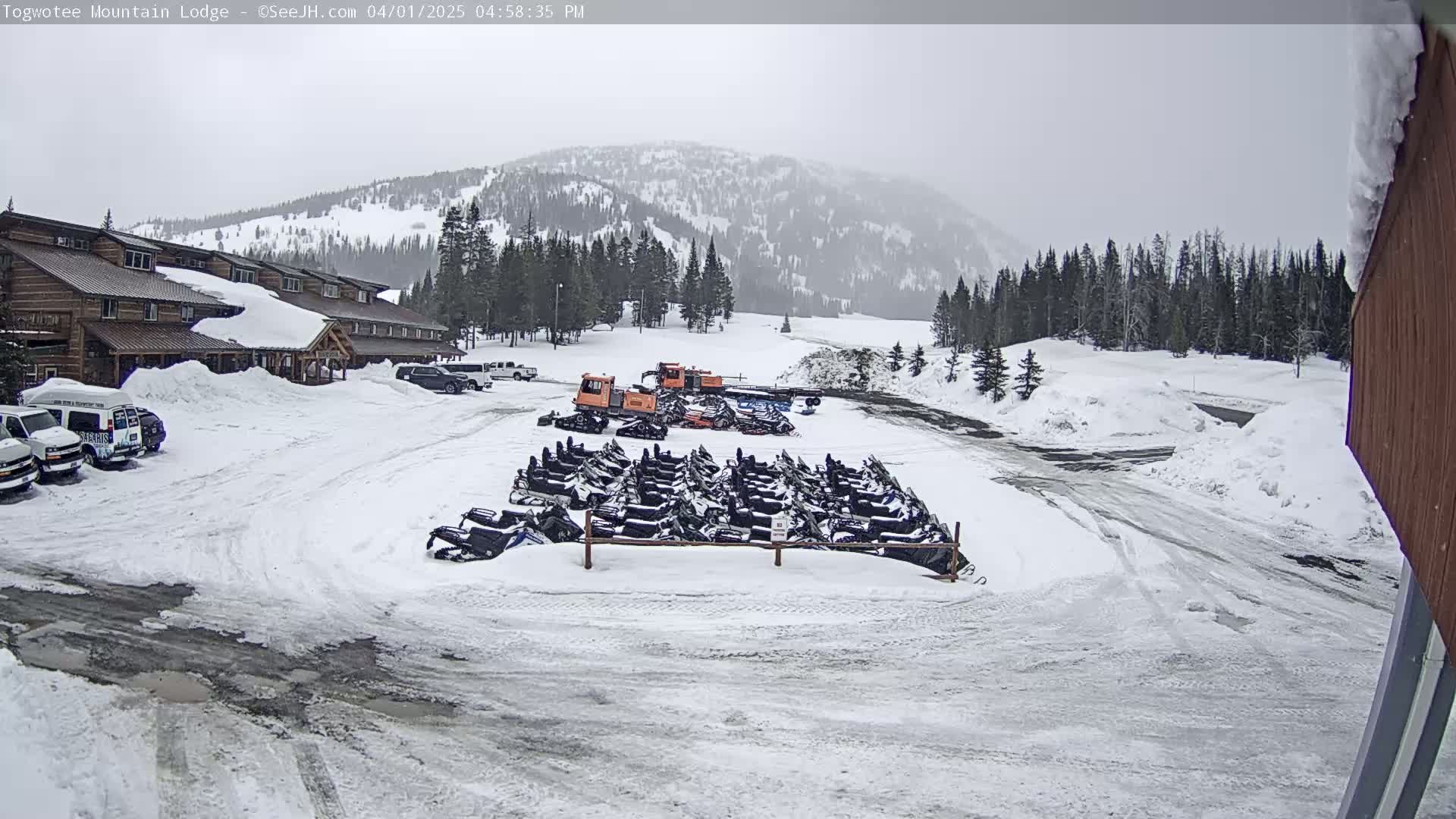 A snow-covered parking lot with a lodge, several snowmobiles, and snowcats is visible in front of a snow-dusted mountain range under an overcast sky.