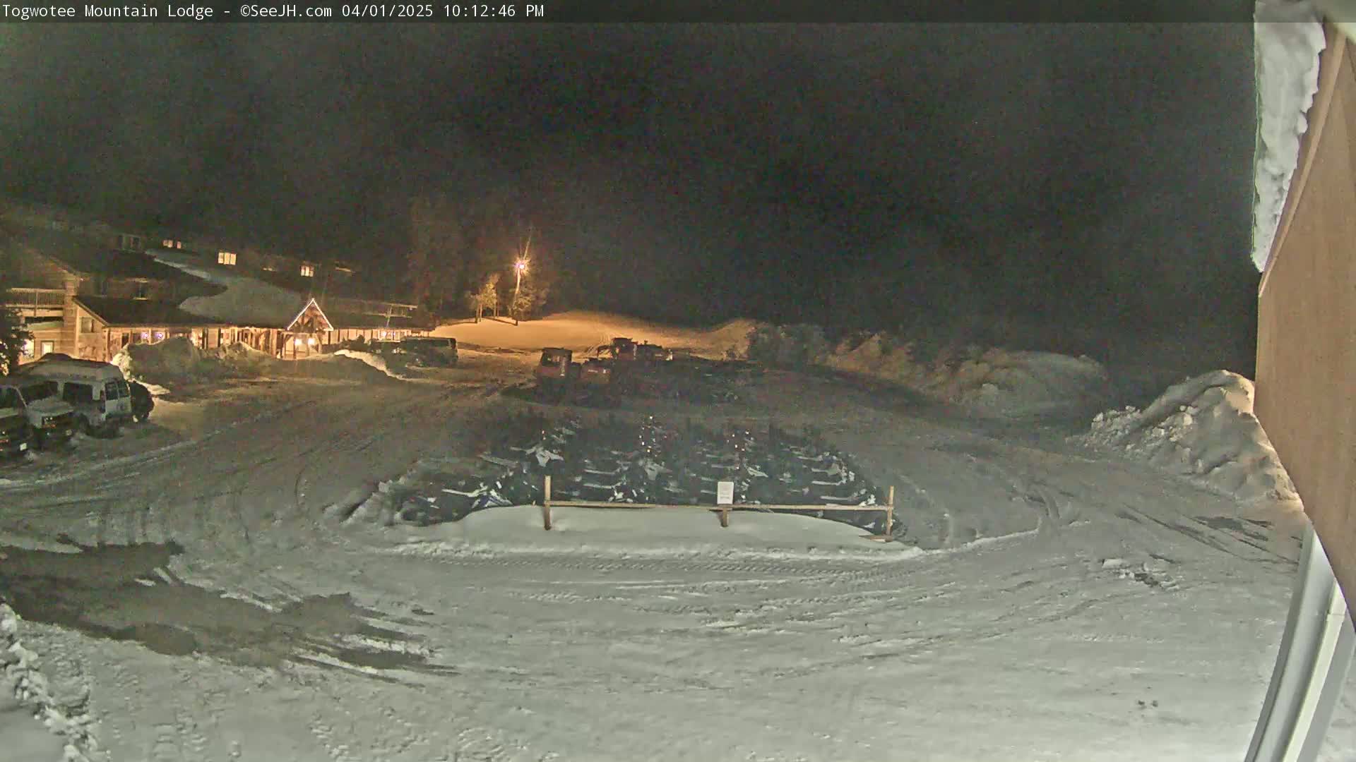 A snow-covered parking lot at night, illuminated by lights, shows a lodge, several vehicles, and numerous snowmobiles parked behind a fence.