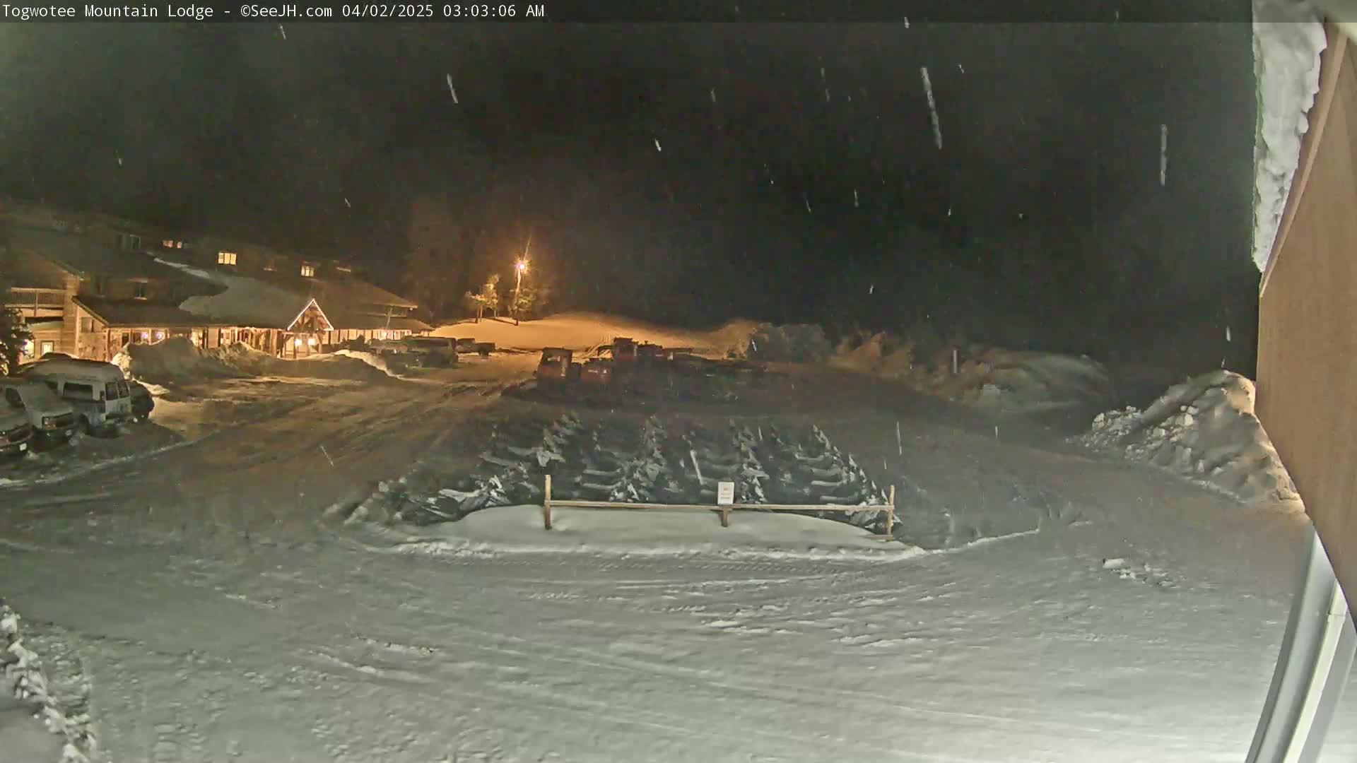 A snow-covered parking lot at night, illuminated by building lights, shows a lodge in the background and several vehicles parked nearby under light snowfall.