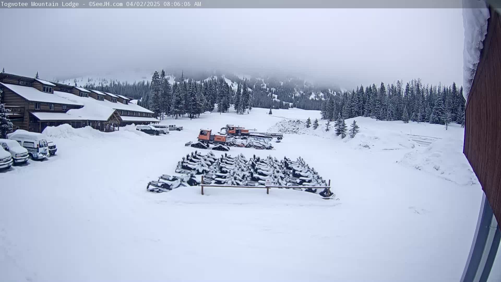 A snow-covered lodge and parking lot with several snowmobiles and snowcats parked outside on an overcast day.