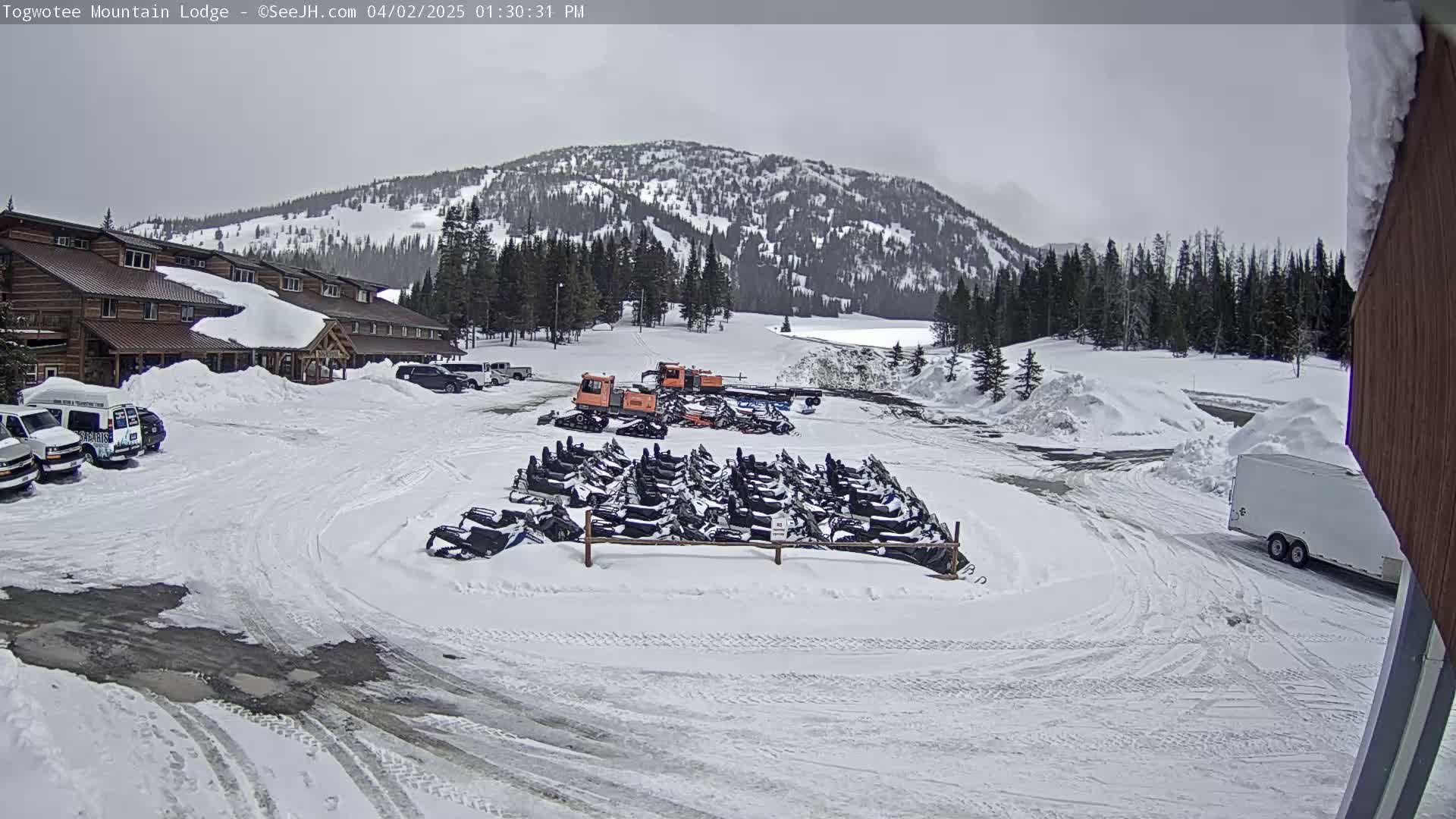 A snow-covered parking lot with numerous snowmobiles parked near a lodge in a mountainous area under an overcast sky.