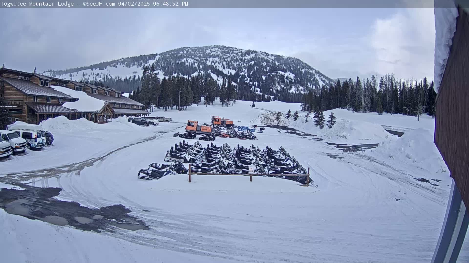 A snow-covered parking lot with numerous snowmobiles parked in front of a lodge, with a snow-covered mountain in the background under a mostly cloudy sky.