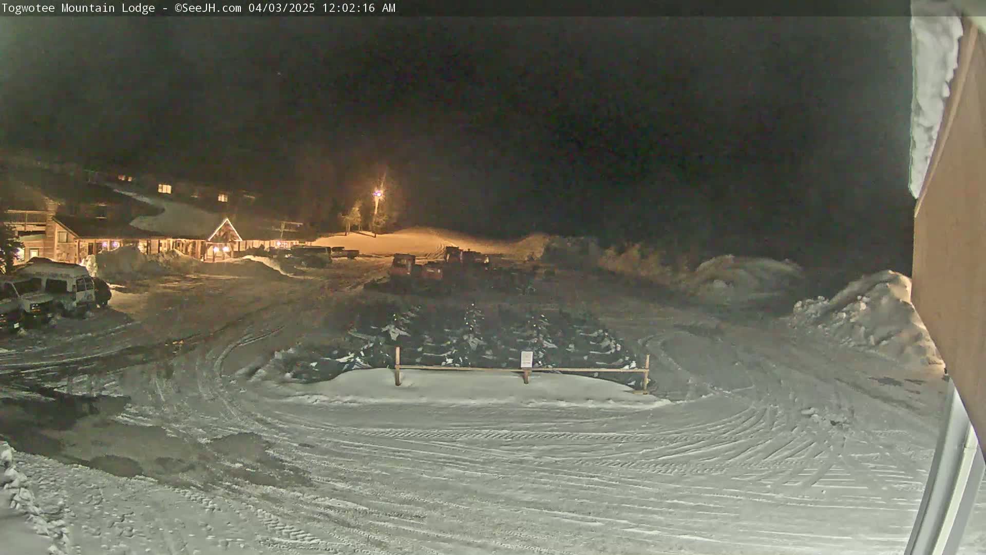 A snow-covered parking lot at night with a lodge in the background, numerous snowmobiles parked, and several vehicles near the building.