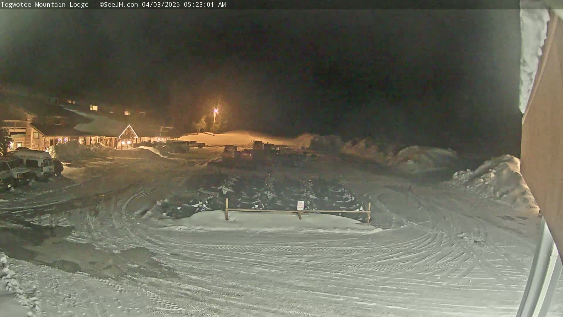 A snow-covered parking lot at night, with a lodge in the background and numerous snowmobiles parked behind a fence.