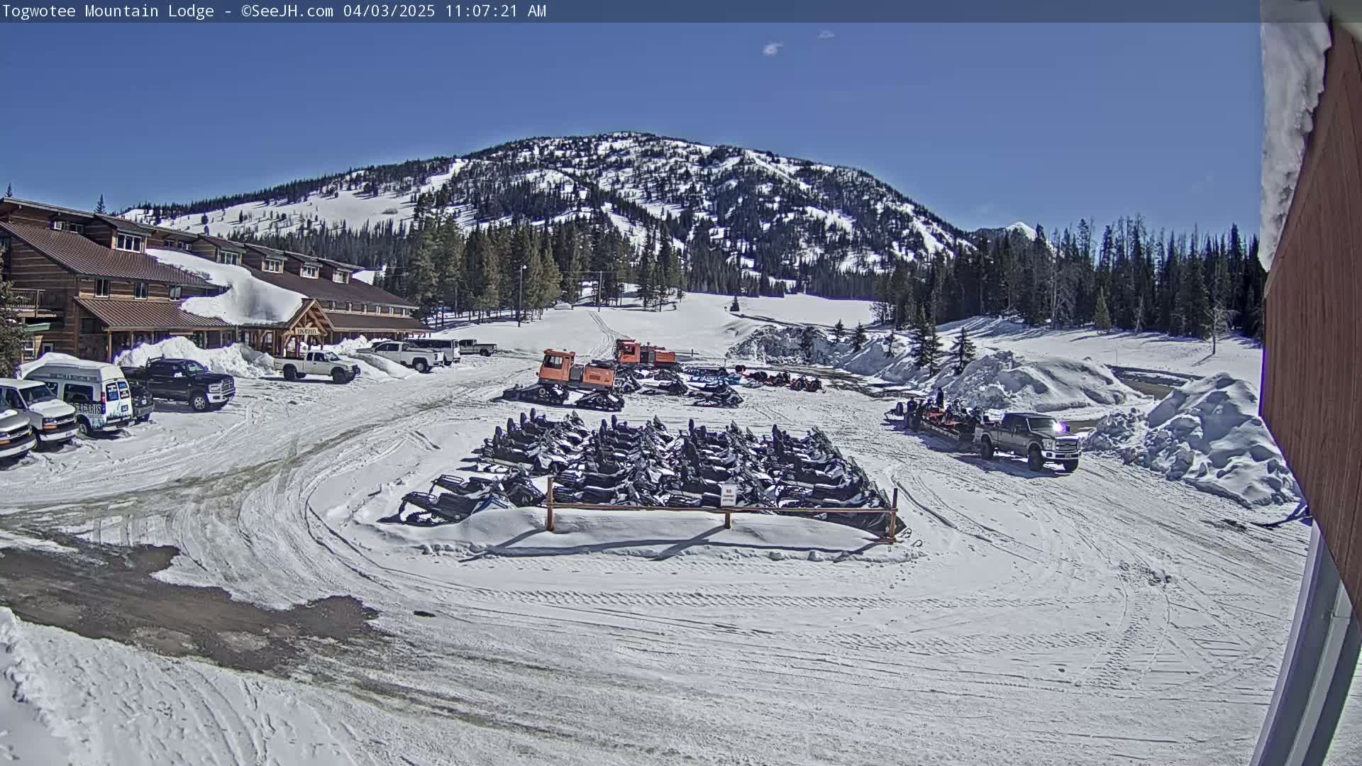 A snow-covered lodge and parking lot with numerous snowmobiles parked outside, under a clear blue sky.