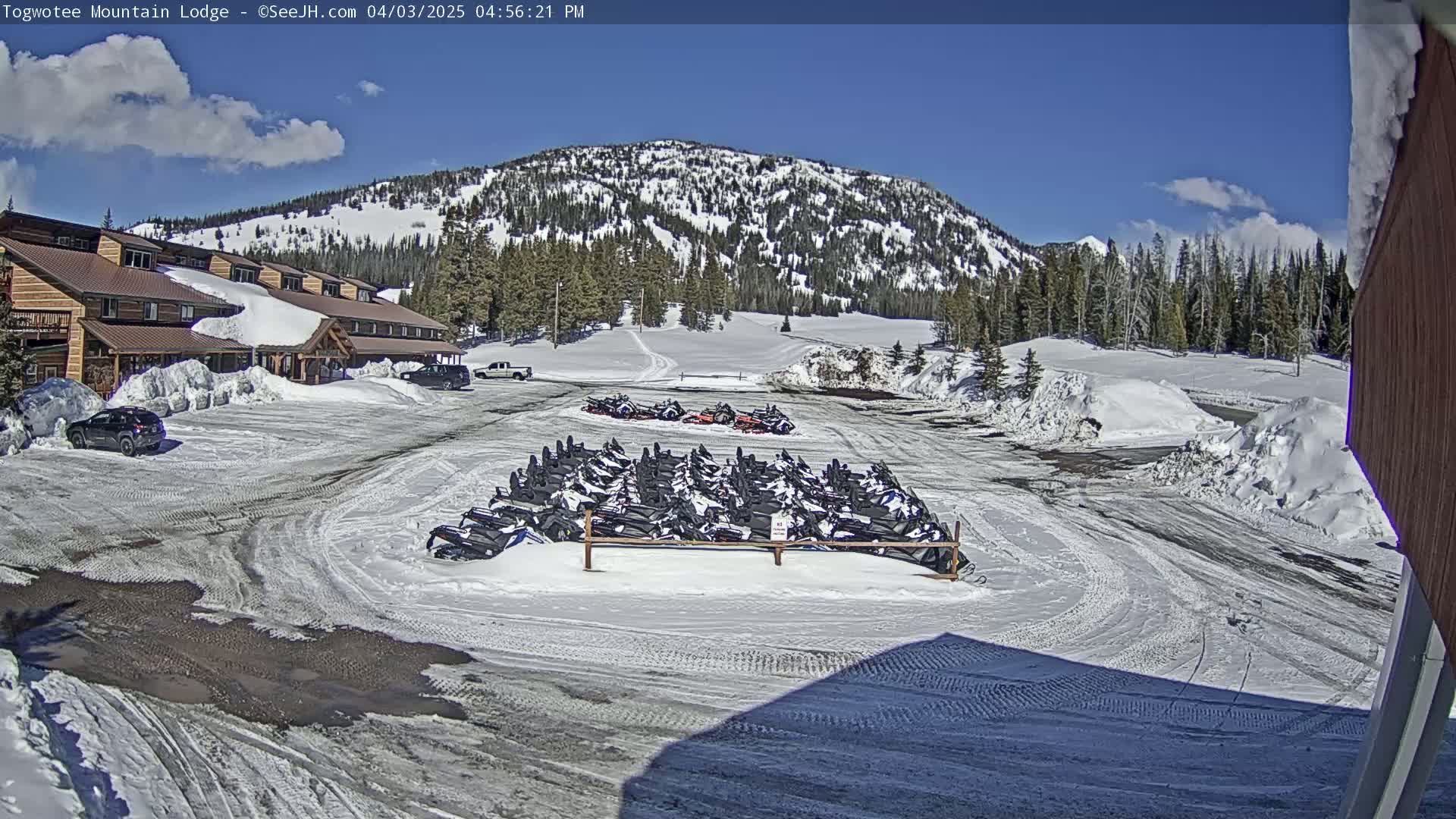 A snow-covered parking lot with numerous snowmobiles parked in front of a lodge, set against a backdrop of a snow-capped mountain range under a sunny sky.