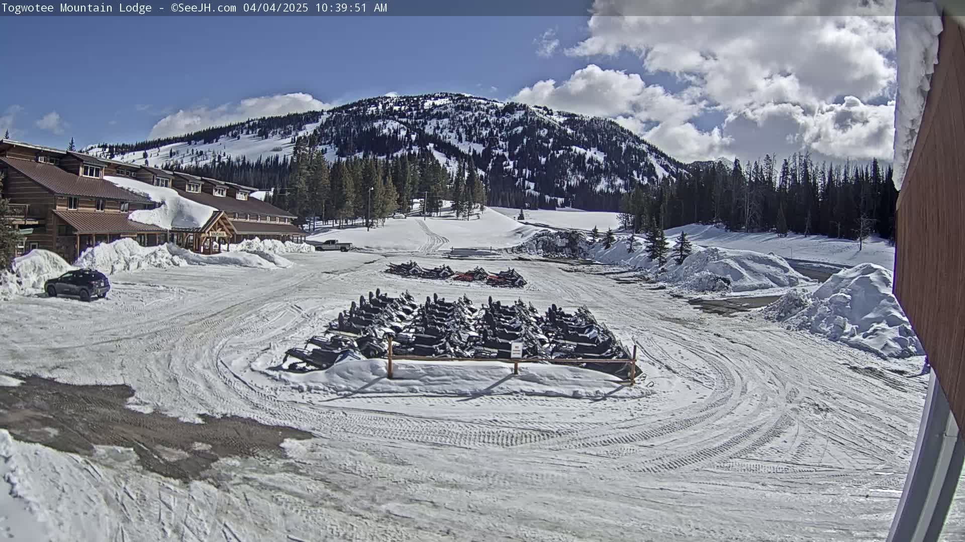A snow-covered parking lot with a lodge and numerous snowmobiles is visible under a partly sunny sky.