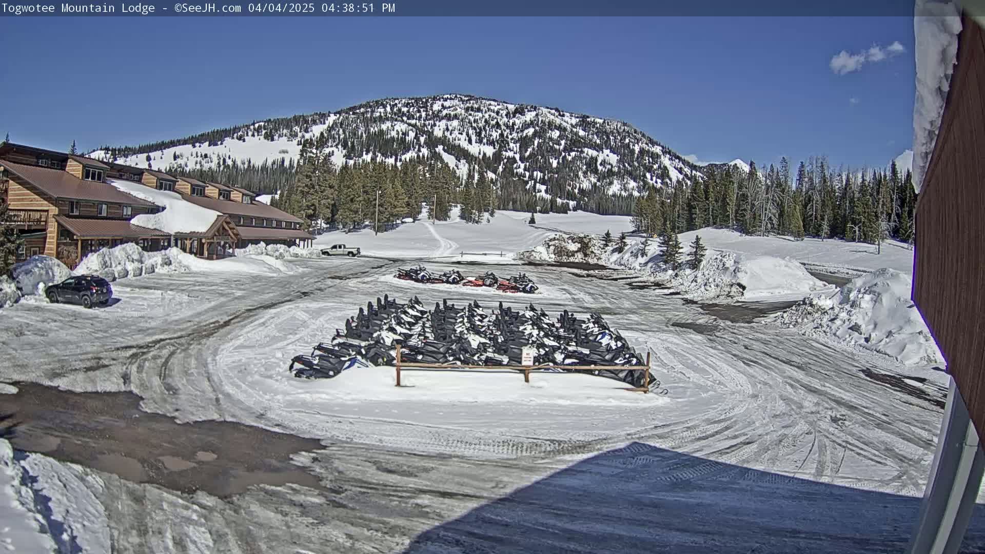 A snow-covered parking lot with numerous snowmobiles parked near a lodge, under a bright, sunny sky, with snow-covered mountains in the background.