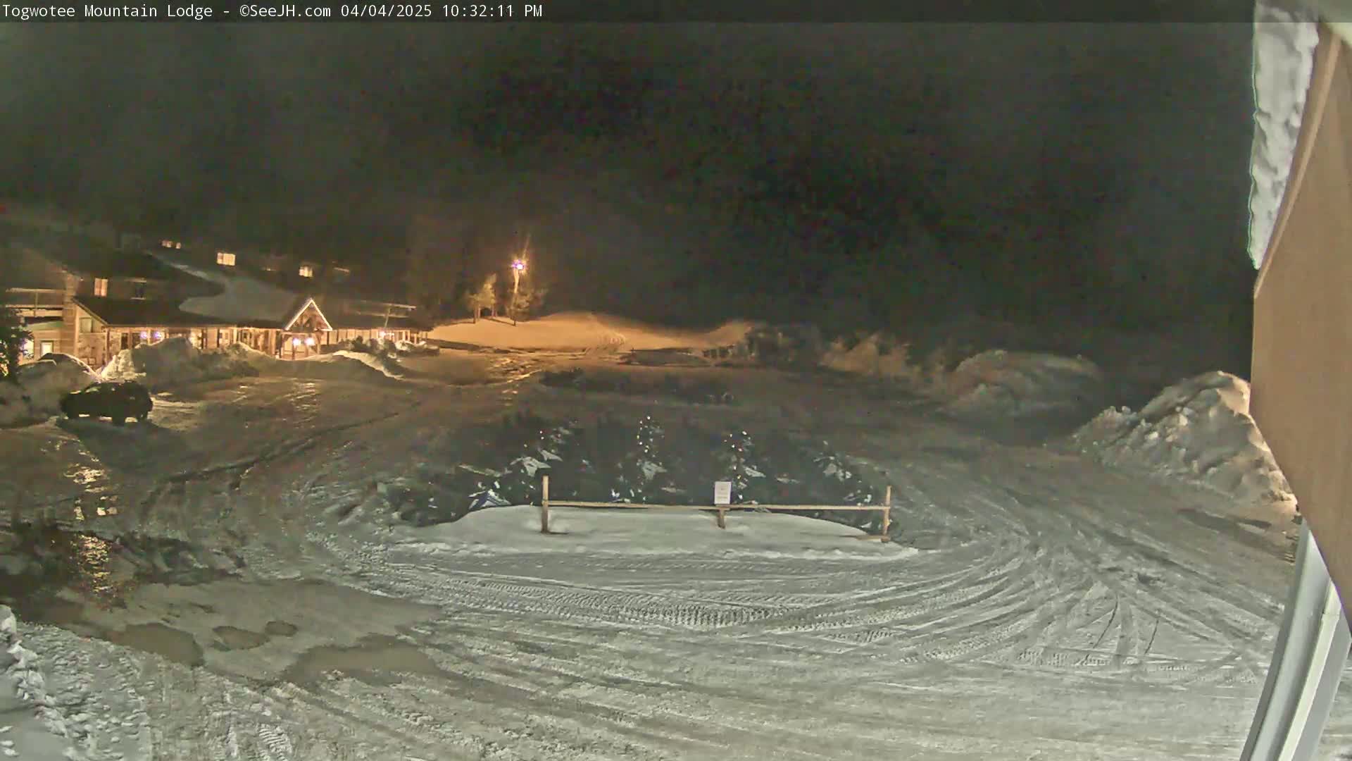A nighttime scene of a snow-covered parking lot with tire tracks and a lodge in the background under a dark sky.