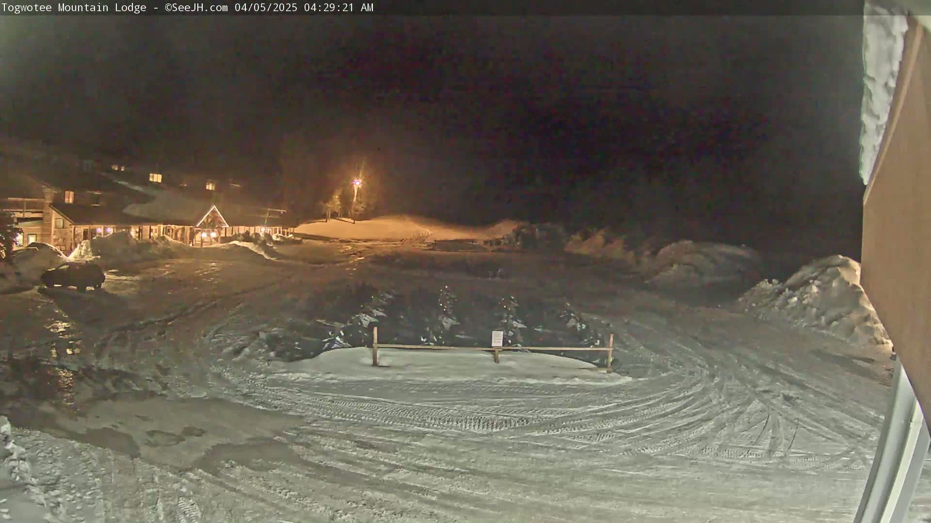 A snow-covered parking lot at night in front of a lit-up lodge, with tire tracks visible on the ground.