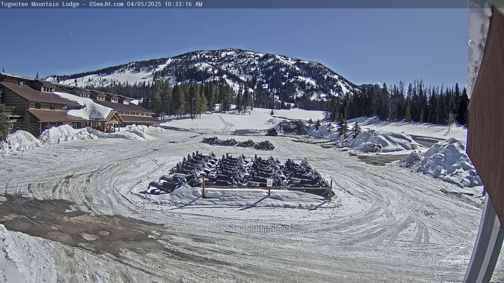 A snow-covered parking lot with numerous snowmobiles parked in front of a lodge, situated against a backdrop of a snow-dusted mountain under a clear, sunny sky.