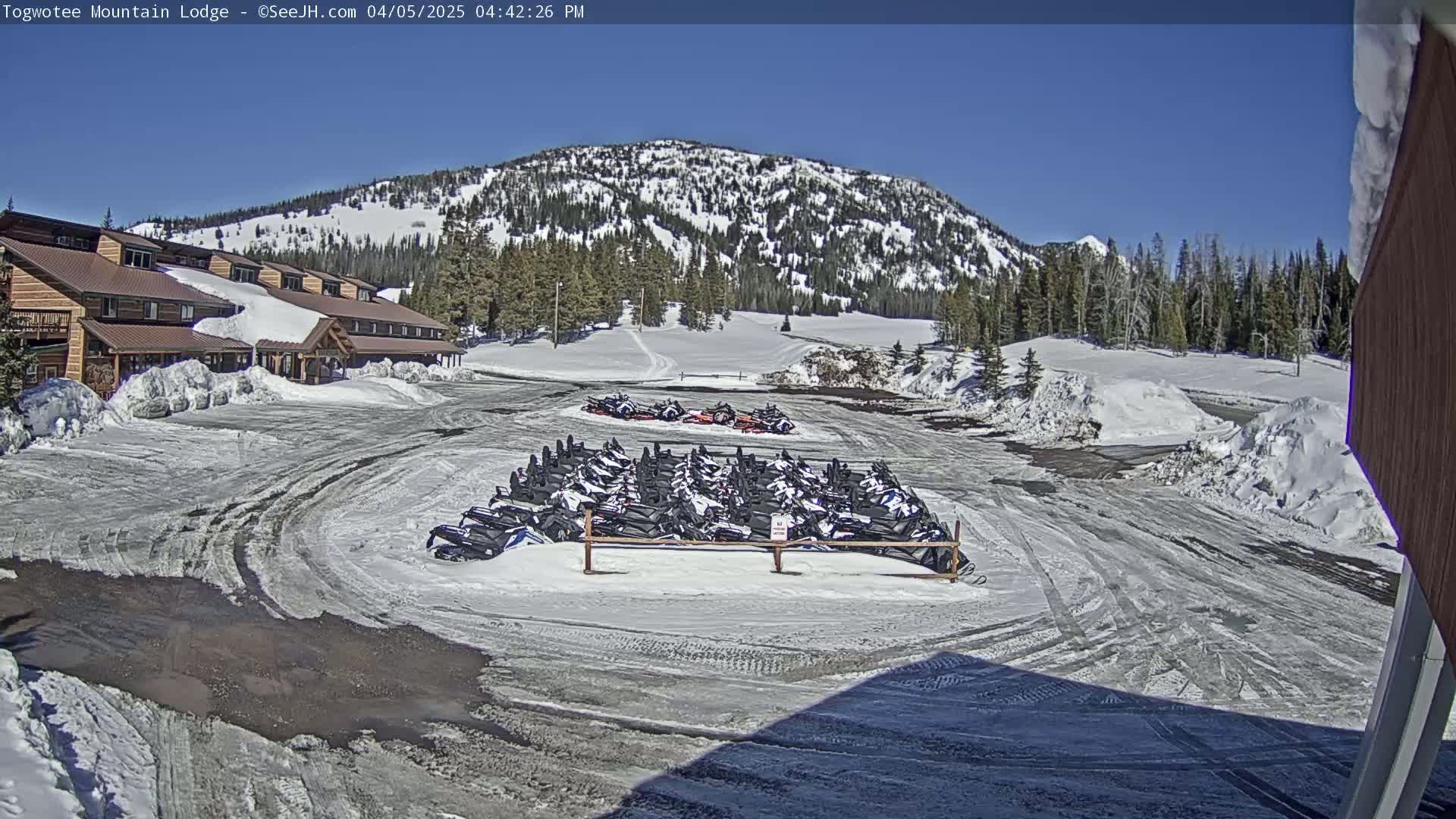 A snow-covered parking lot with numerous snowmobiles parked near a lodge, under a bright sunny sky with a snow-capped mountain in the background.