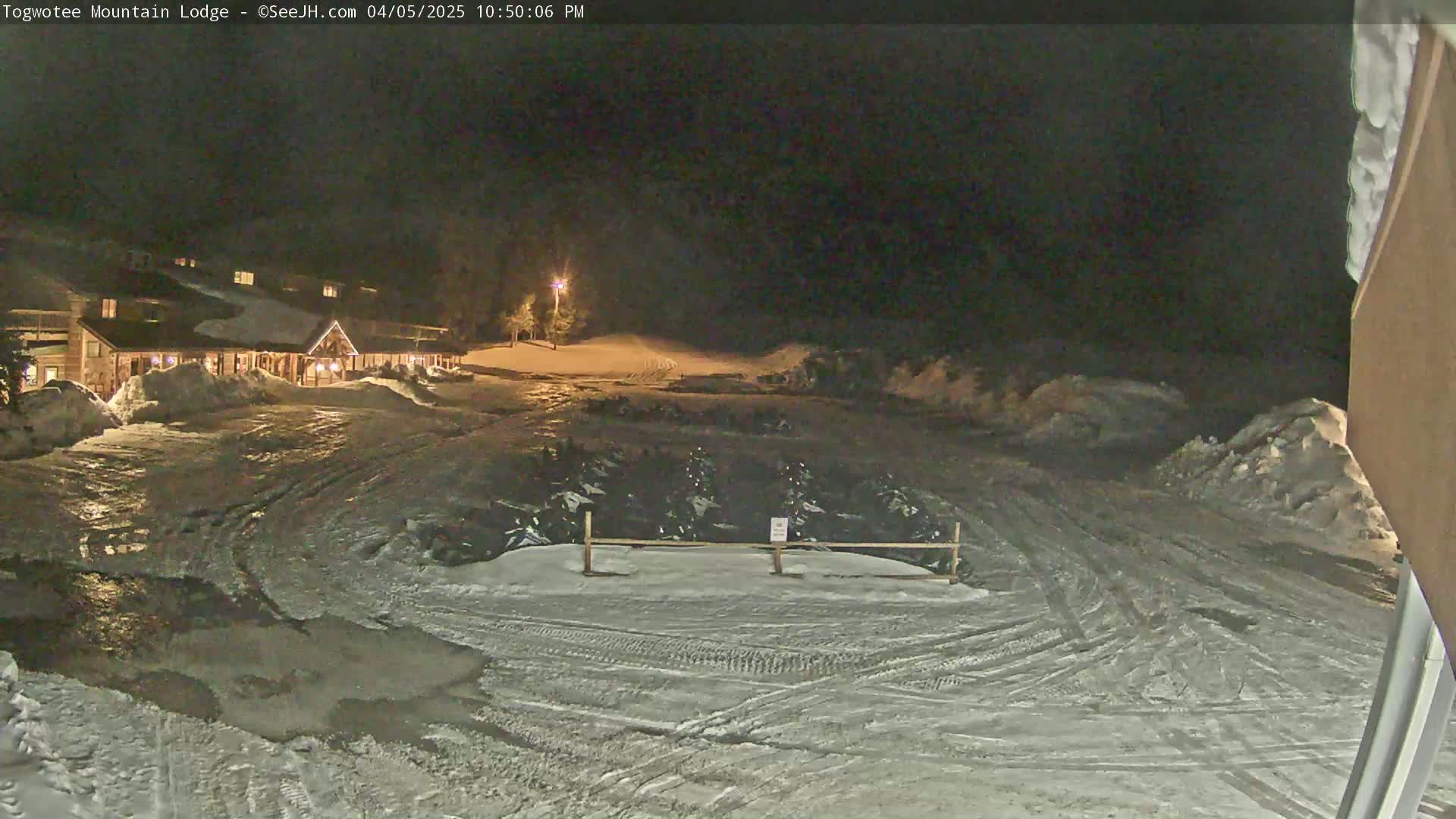 A snow-covered parking lot at night in front of a lit-up lodge, with tire tracks visible on the ground.
