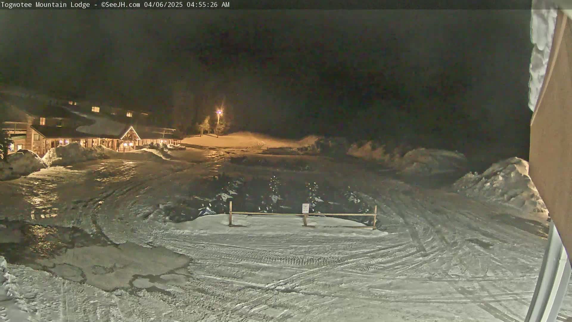 A snow-covered parking lot at night, in front of a lit lodge, shows tire tracks in the snow and some vehicles parked behind a fence.