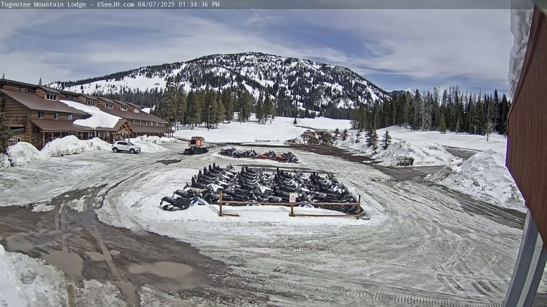 A snow-covered parking lot with numerous snowmobiles parked near a lodge and a snow-covered mountain in the background under a partly cloudy sky.