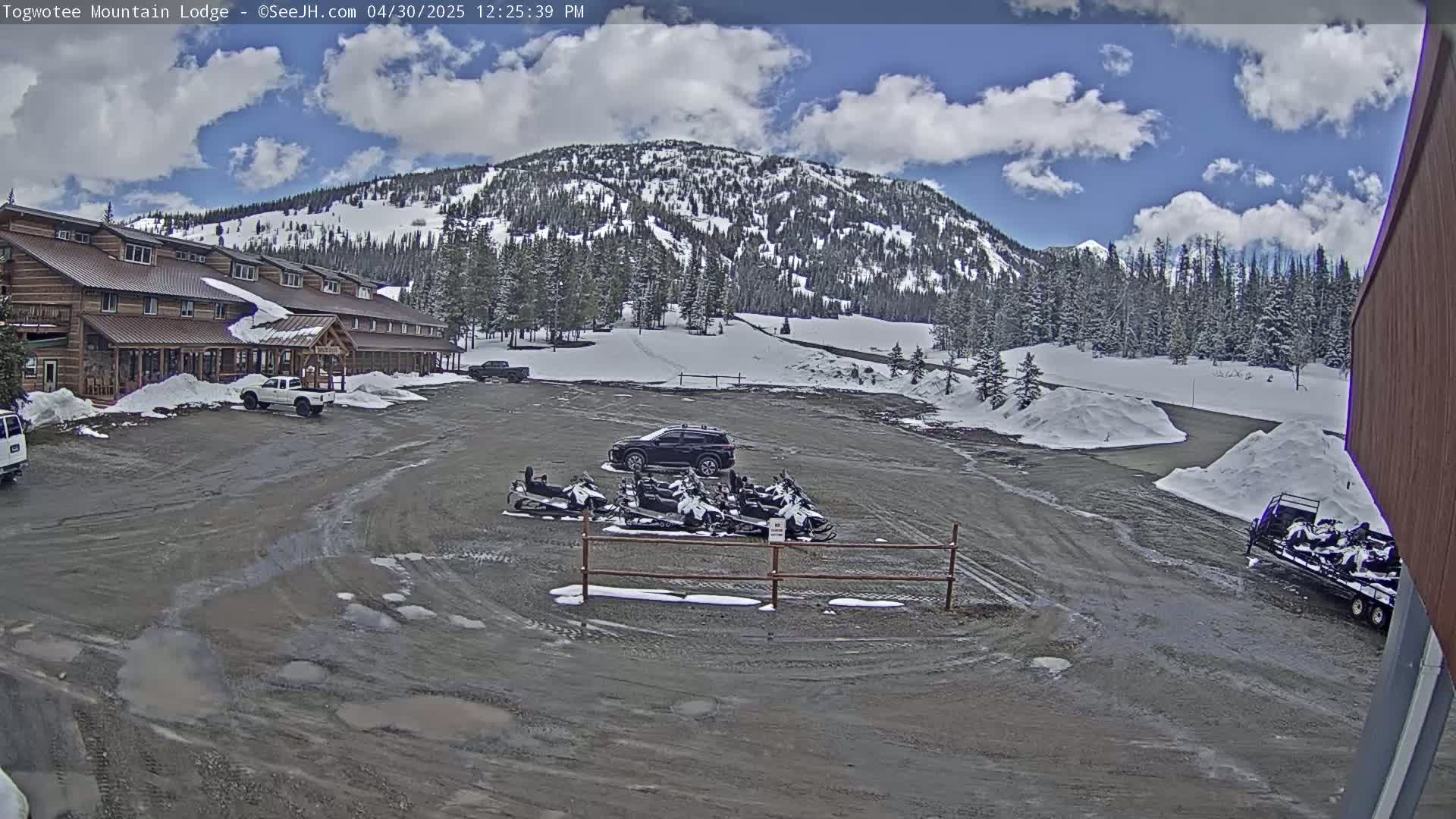 A large, rustic lodge sits in a parking lot with several snowmobiles and a dark-colored SUV, all under a partly cloudy sky, with snow on the ground and nearby mountainside.
