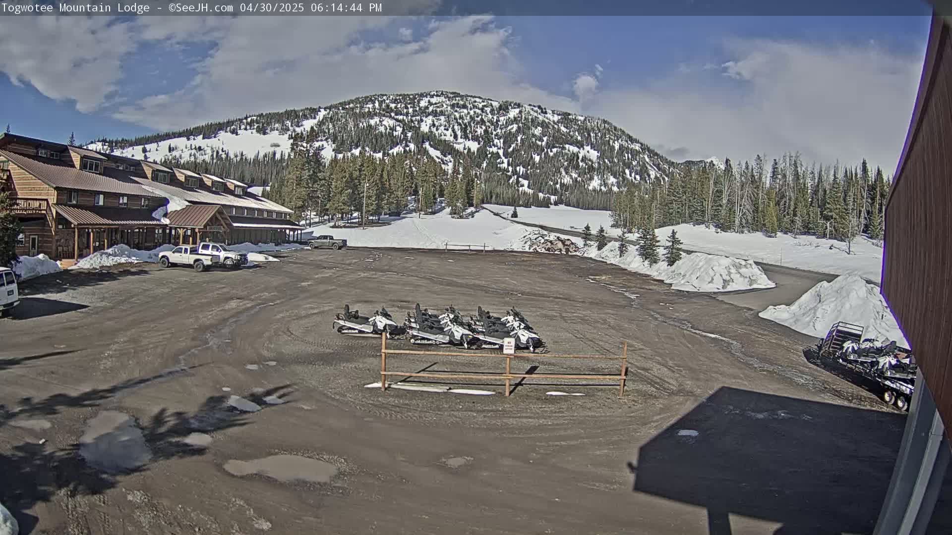 A lodge sits in a parking lot with several snowmobiles, partially snow-covered ground, and a mountain in the background under a partly sunny sky.
