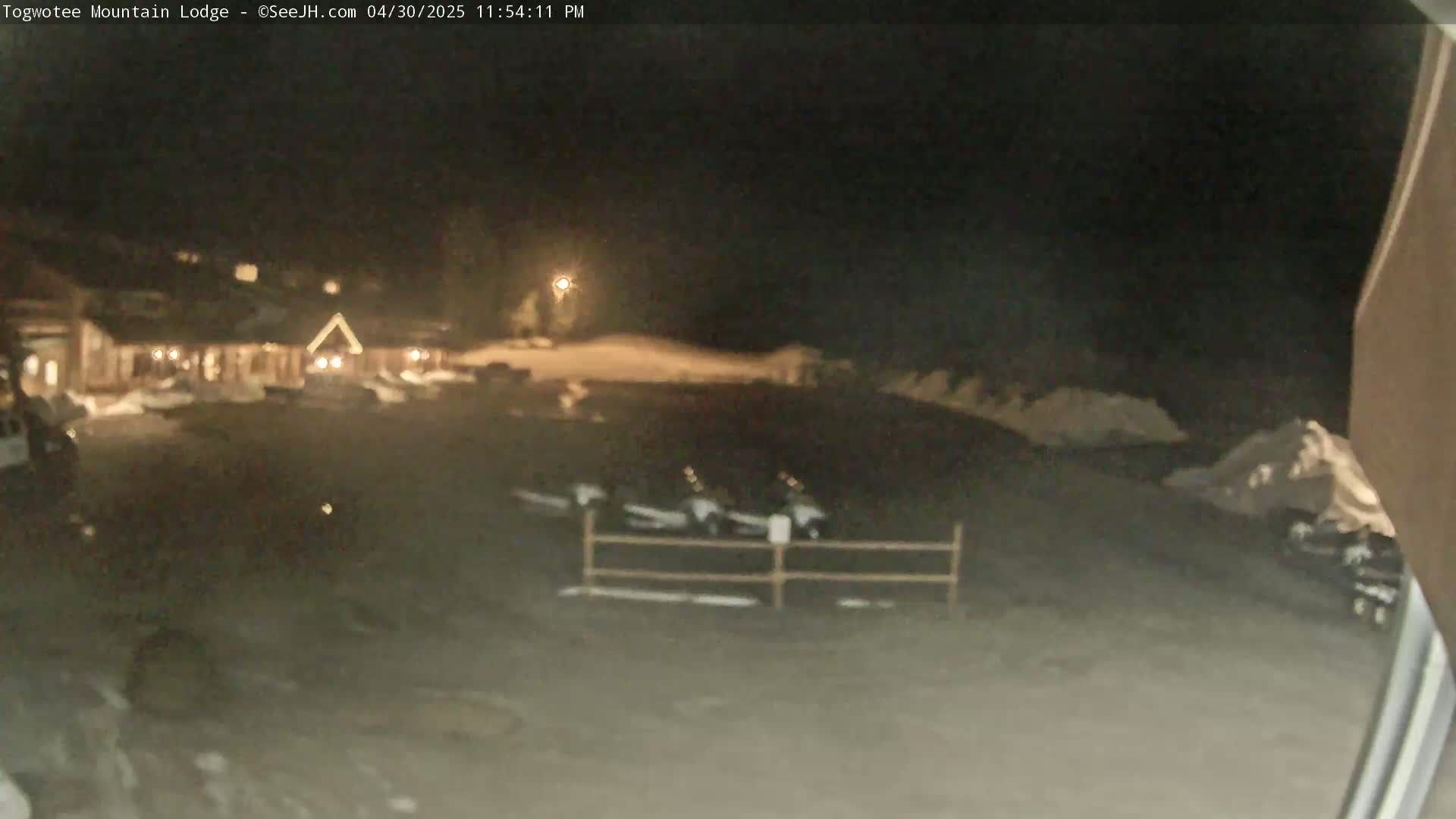 A nighttime, snowy scene shows a lodge, a parking lot with several vehicles, and a low fence.