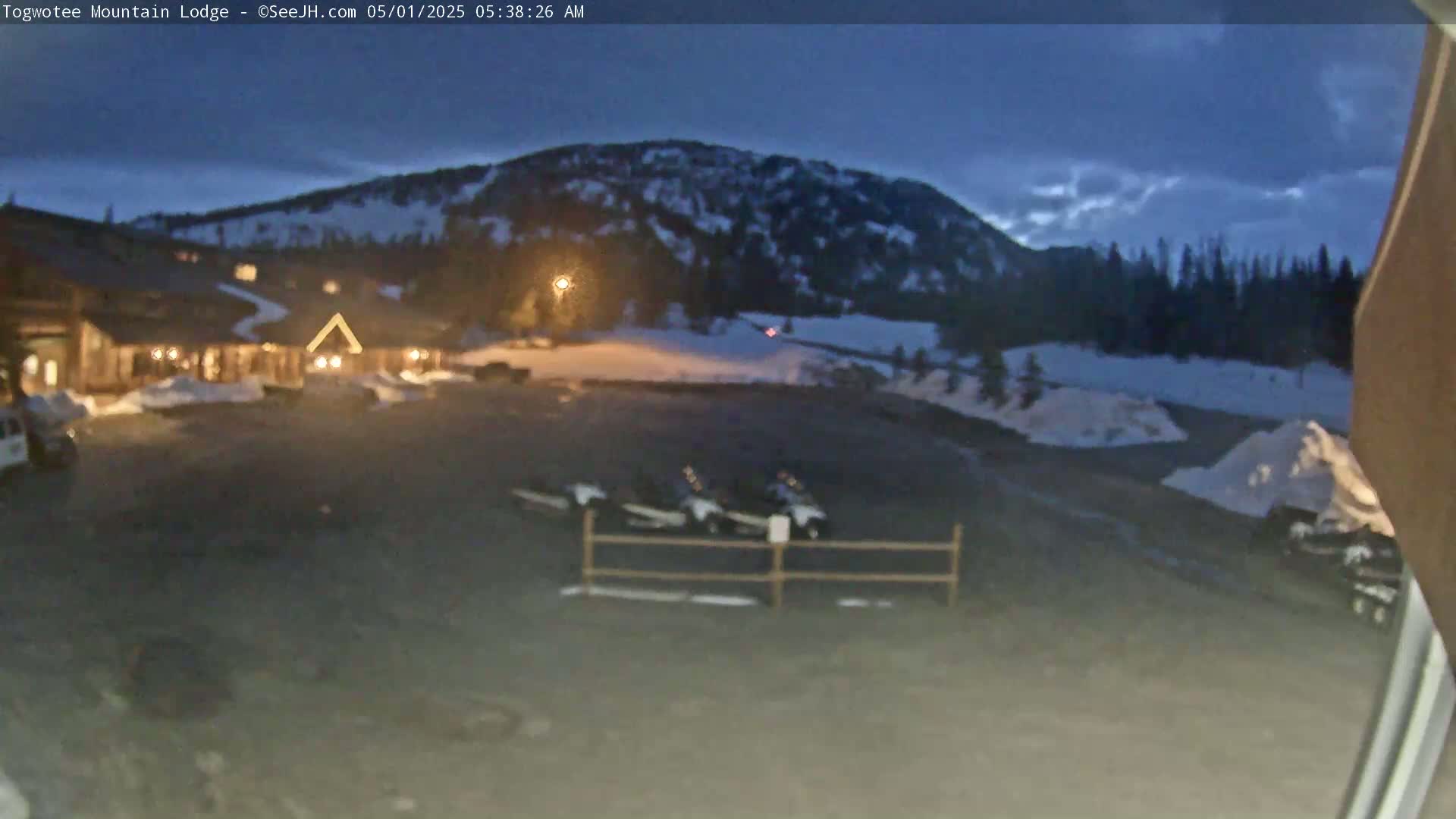 A nighttime view of a lodge parking lot with snow on the ground, several vehicles parked, and a mountain in the background under a dark, partly cloudy sky.