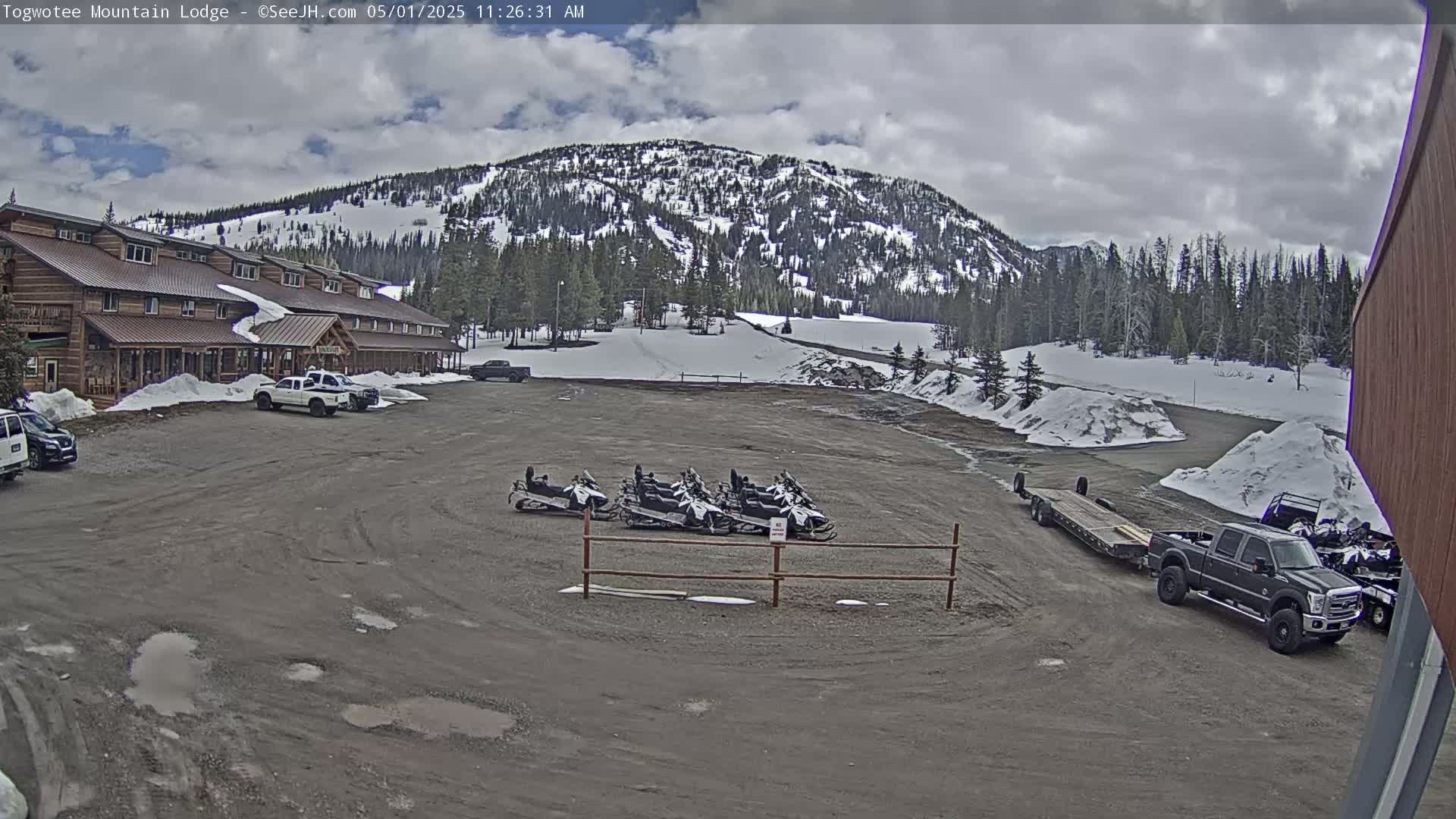 A large, wood-sided lodge sits in a parking lot with several snowmobiles and a pickup truck towing a trailer, all under a partly cloudy sky, with snow on the ground and in the surrounding hills.