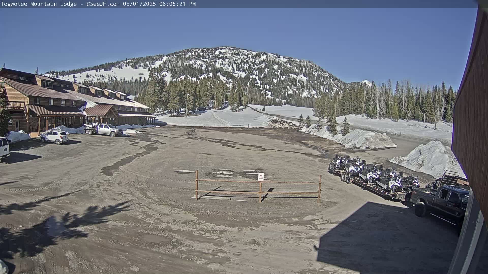 A lodge with a parking lot in front, several vehicles, and snowmobiles on a trailer are situated in front of a snow-covered mountain under a clear blue sky.