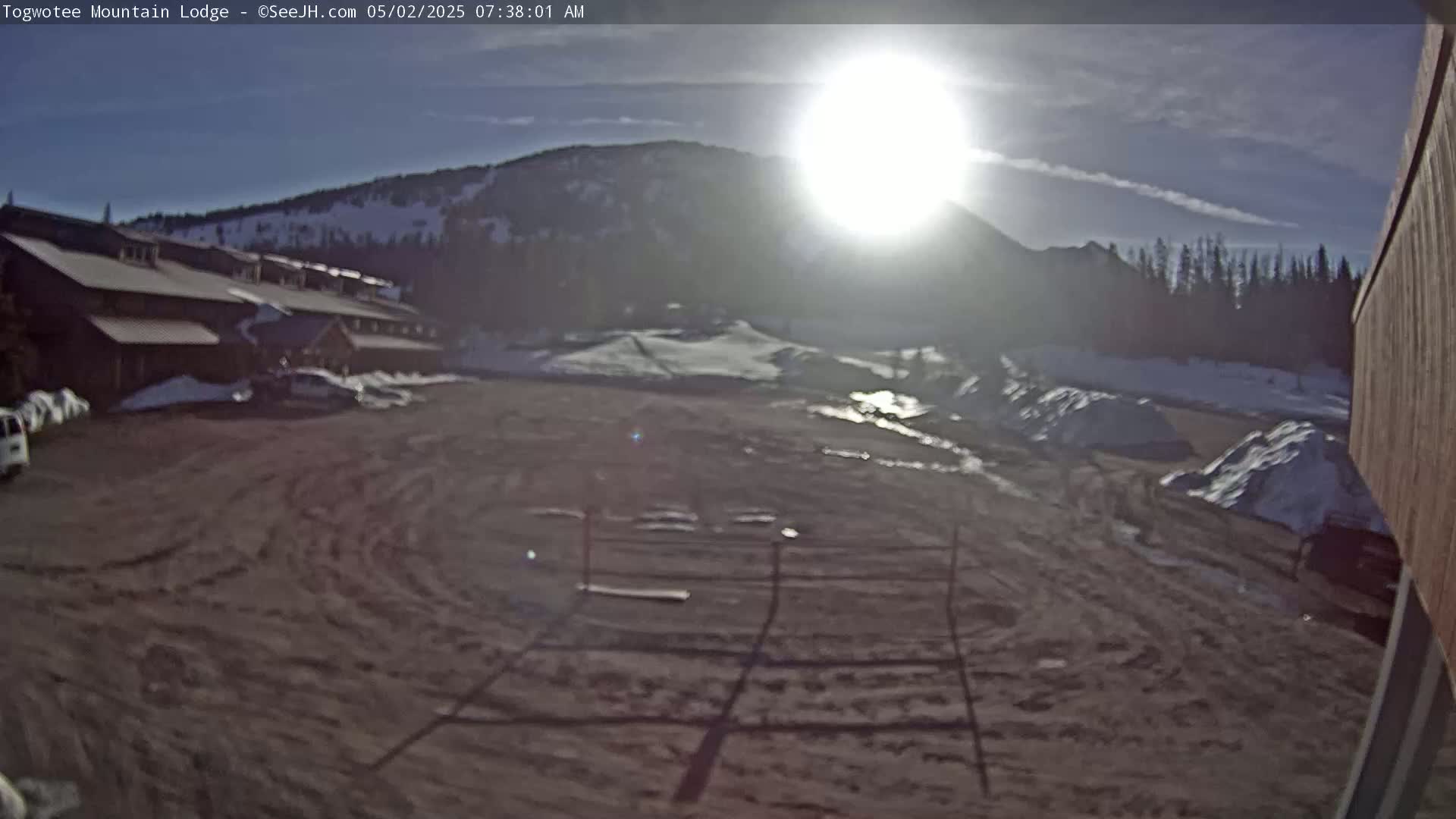 A mostly sunny morning reveals a lodge, parking lot, and snowy hillside in a mountain setting.