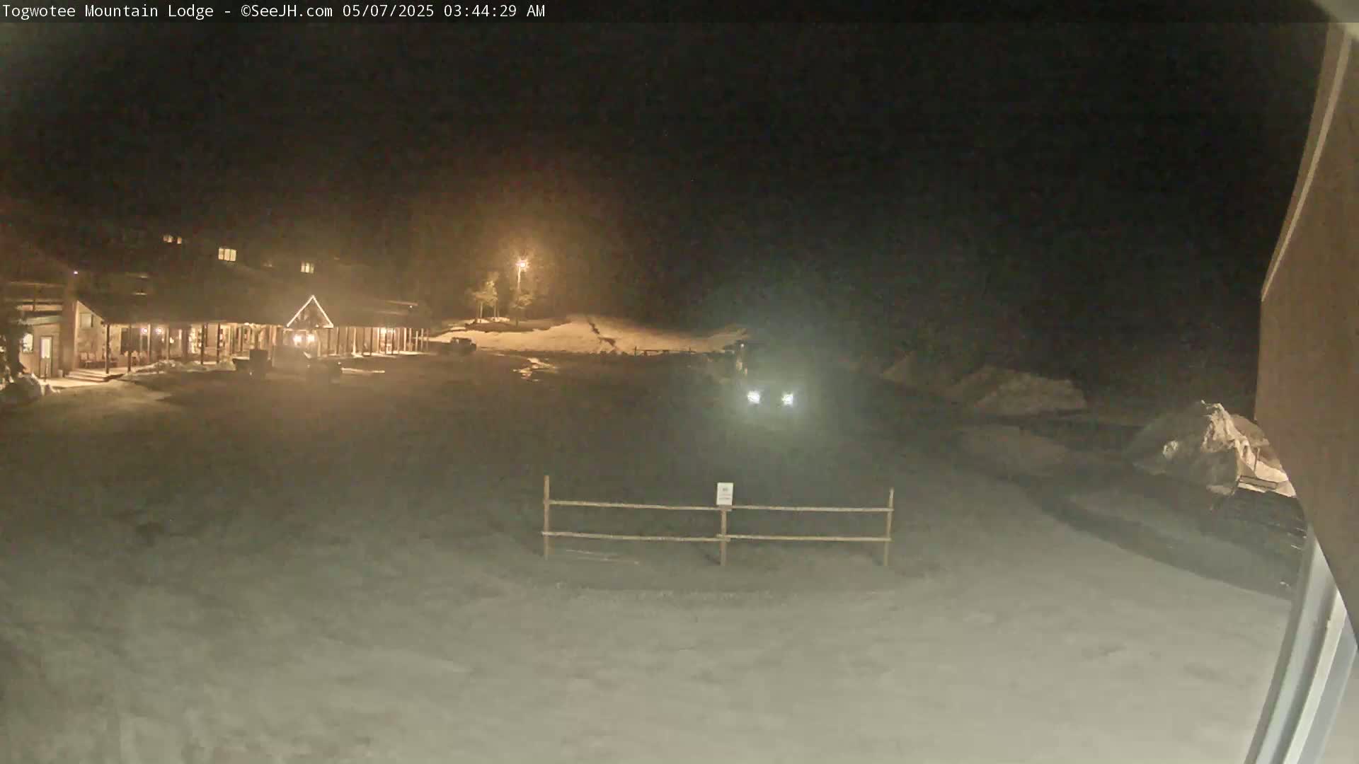 A lodge is illuminated at night in a snow-covered area, with vehicles visible in the parking lot and headlights from a distant vehicle.