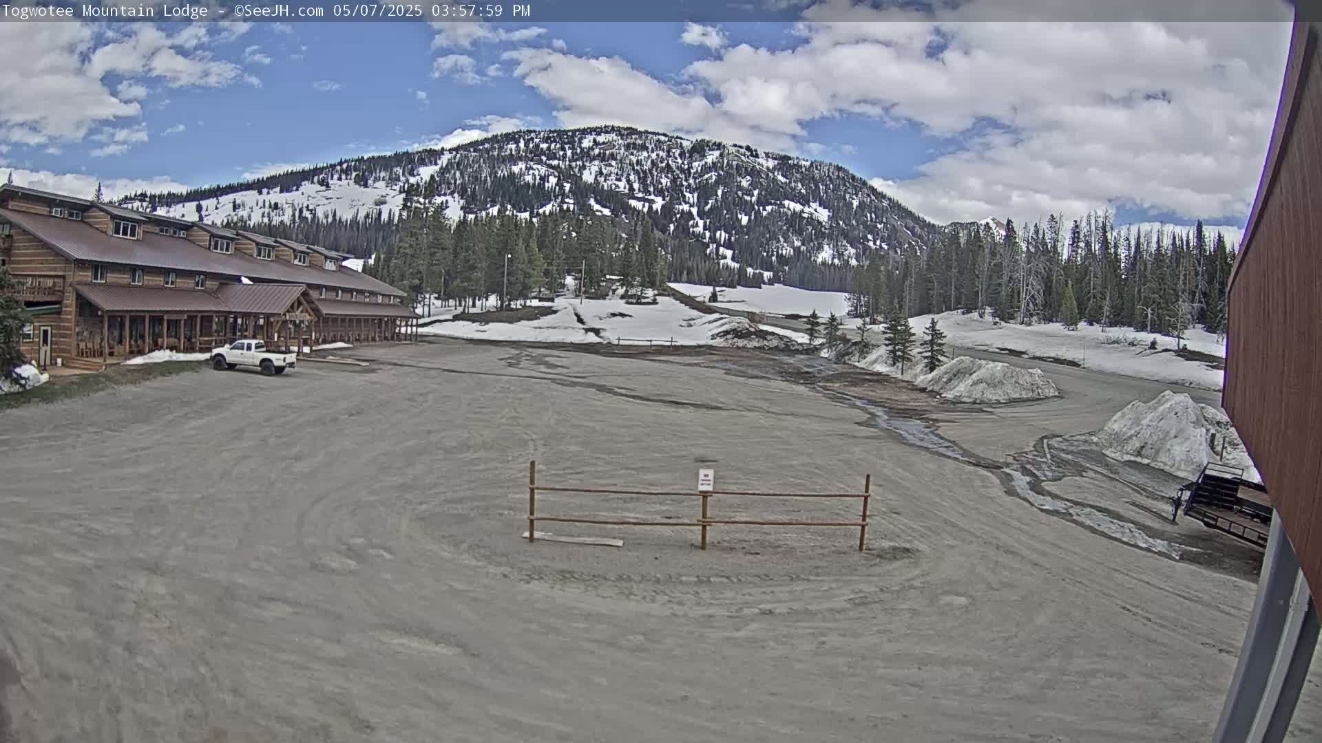 A large, wood-framed lodge sits in a mostly-empty parking lot with patchy snow and melting snow piles, in front of a mountain range with some snow on its peaks under a partly cloudy sky.