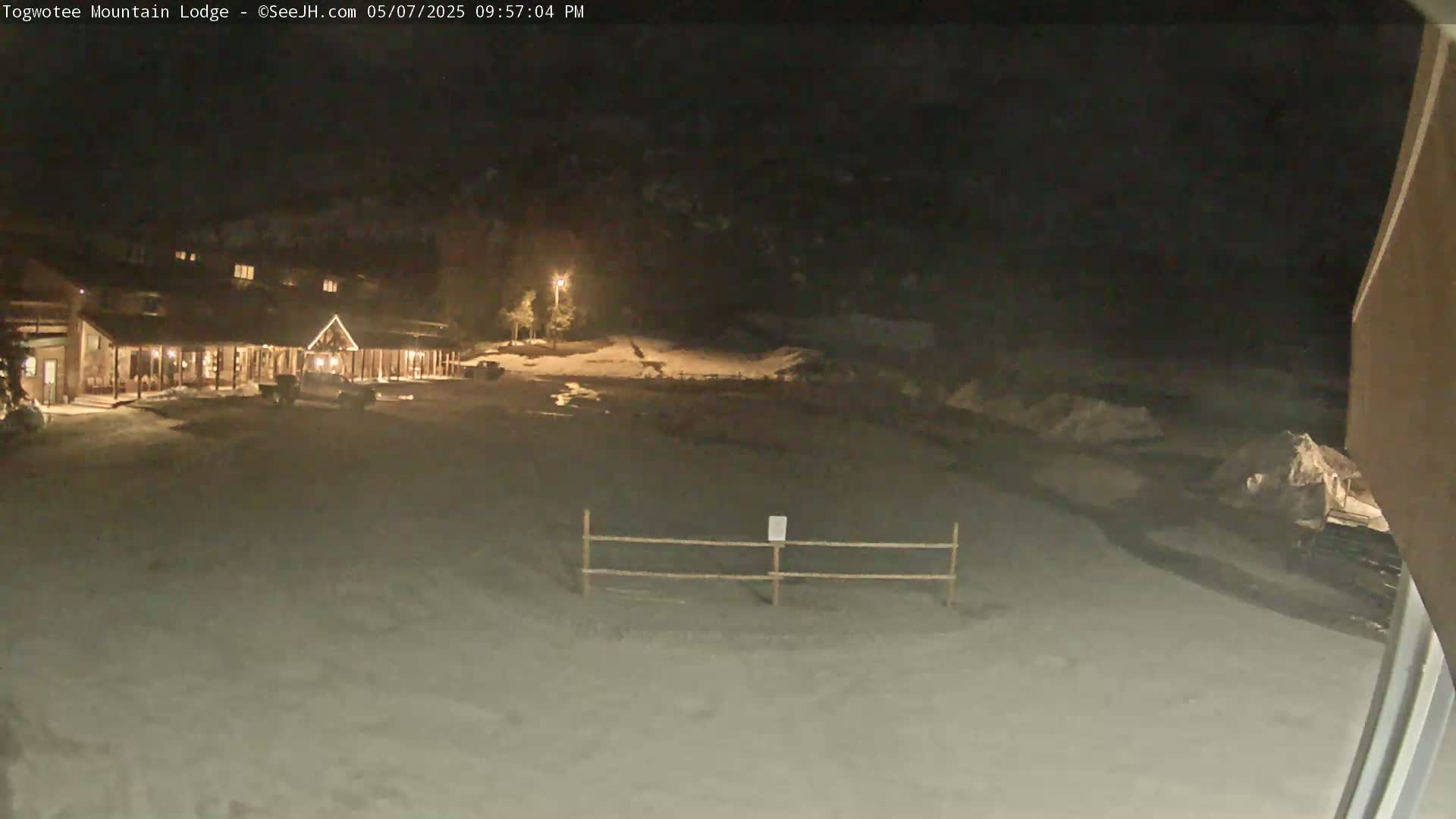 A snow-covered nighttime scene shows a lodge lit up with some vehicles parked in front, a low wooden fence in the foreground, and a dark, snow-dusted landscape beyond.