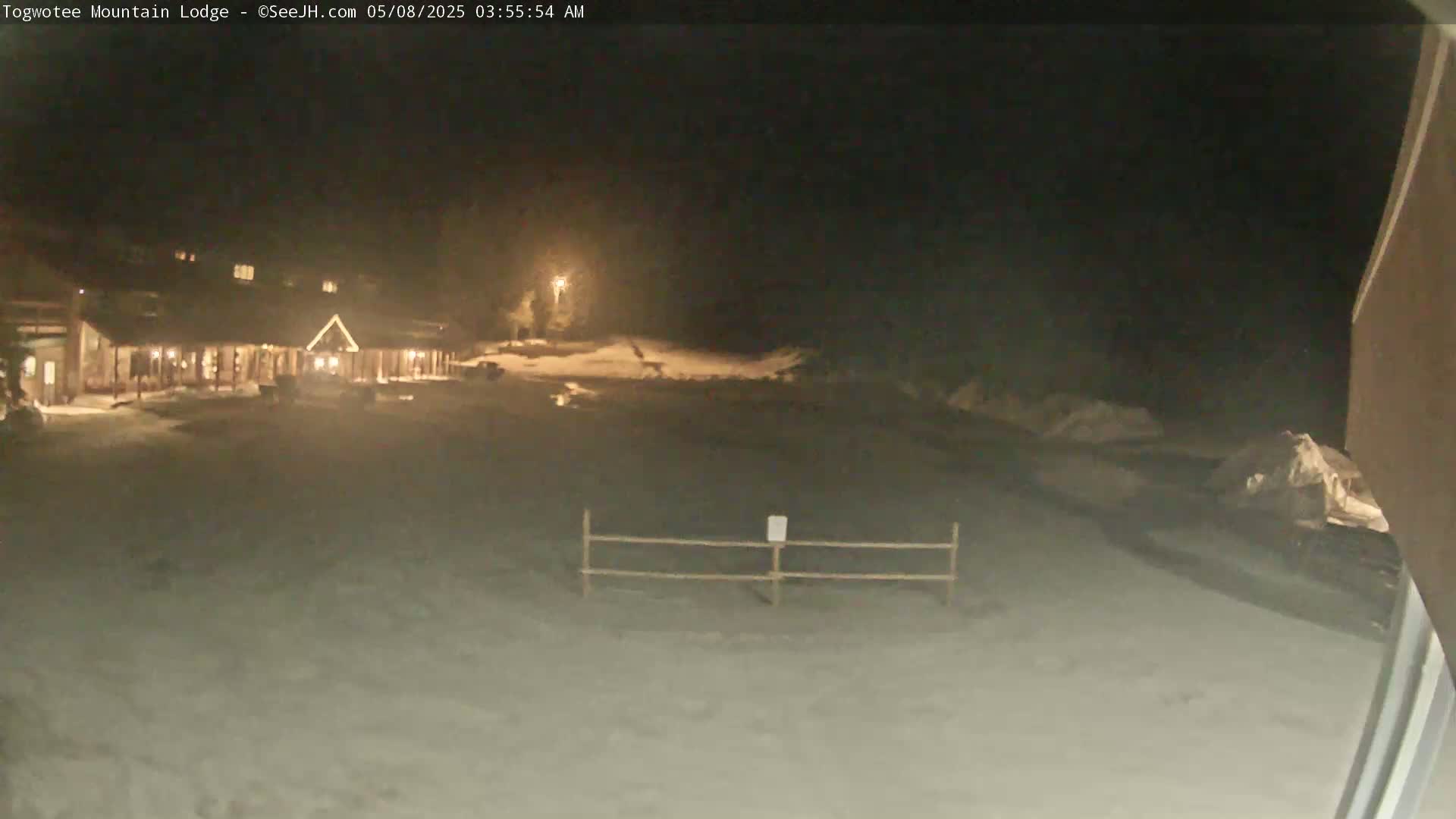 A snow-covered area at night shows a lodge lit up in the distance, with a short fence in the foreground.