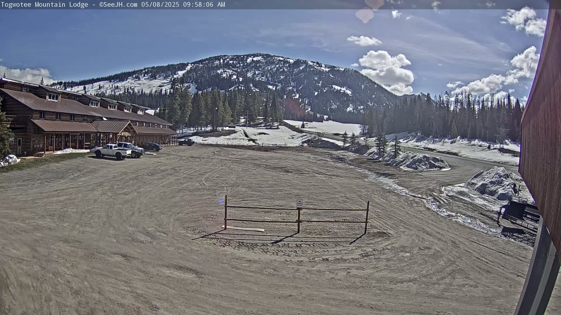 A lodge sits in a mostly empty parking lot, with snow-dusted mountains and a partly cloudy sky visible in the background.