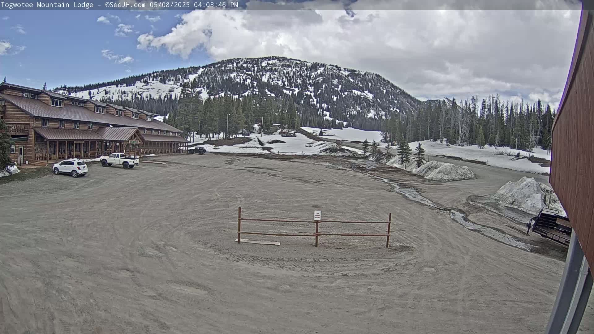 A large lodge sits near a mostly empty parking lot in front of a snow-dusted mountain under a partly cloudy sky.
