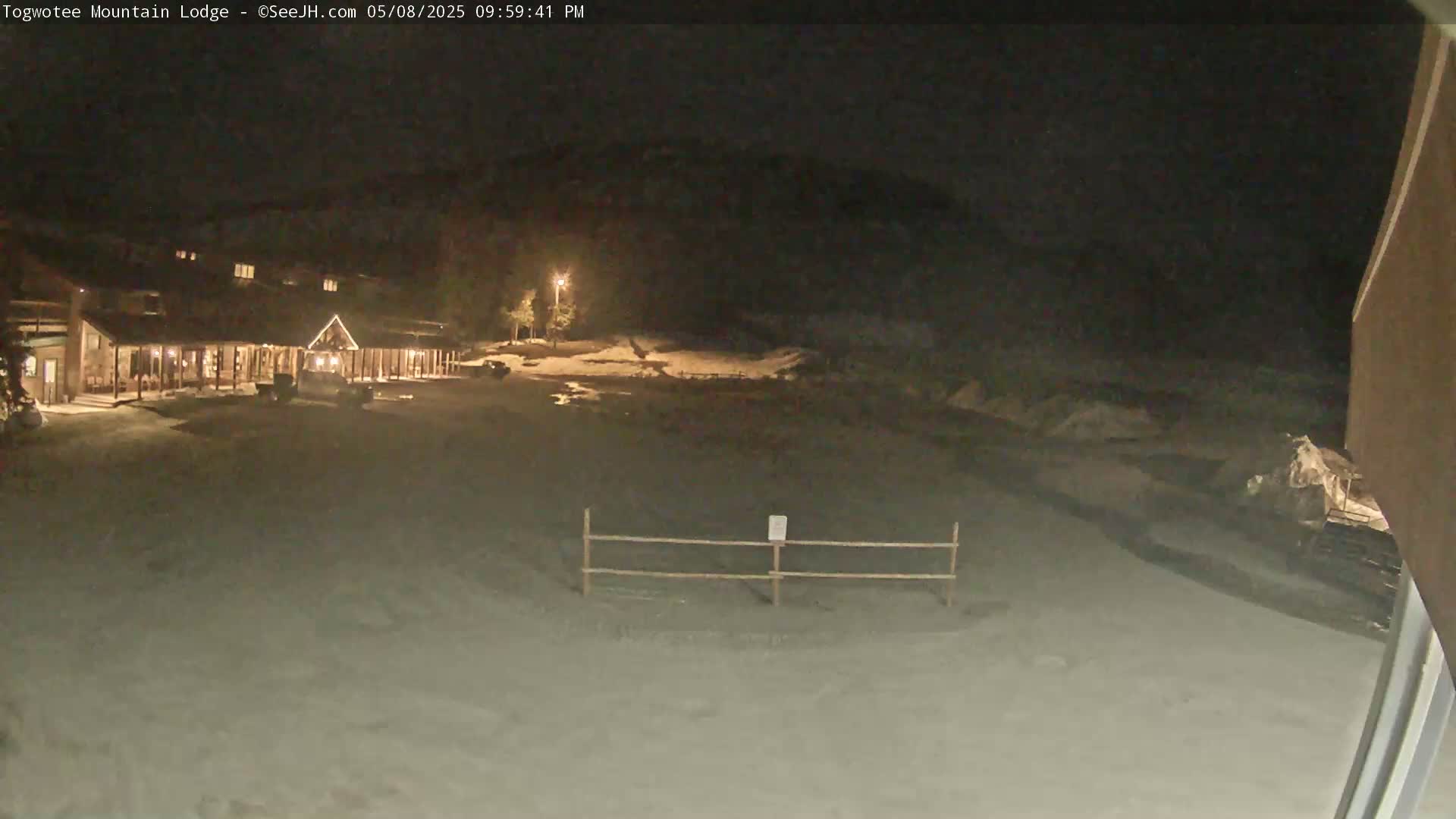 A nighttime view of a snow-covered lodge parking lot and surrounding area, illuminated by artificial lights.