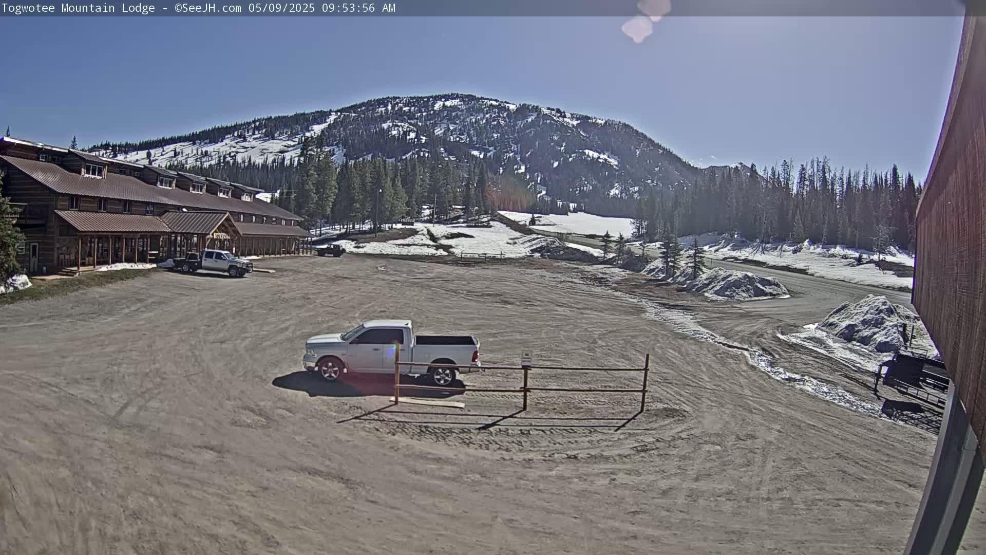 A large, rustic lodge is situated in a parking lot with a few vehicles, overlooking a snowy mountain landscape under a sunny sky.