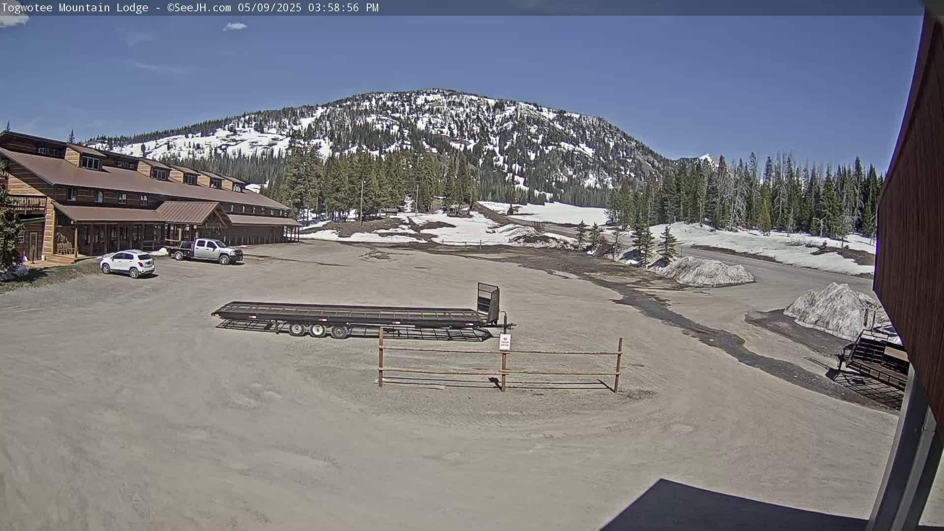 A large lodge with a gravel parking lot in front sits below a snow-dusted mountain range under a clear blue sky.