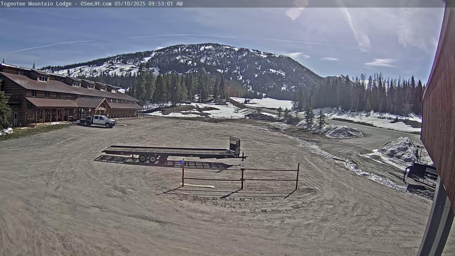 A lodge sits in a parking lot with a flatbed trailer nearby, overlooking a mountain range with patches of snow under a mostly sunny sky.