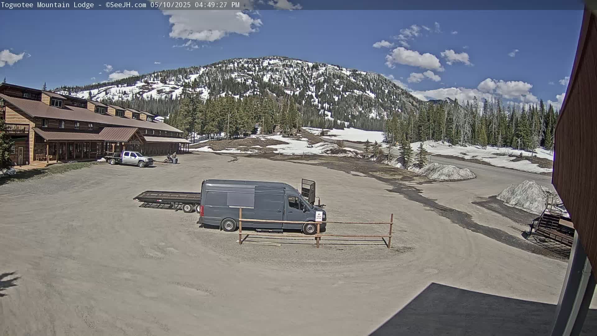 A lodge with a parking lot containing a van and truck is situated in front of a mountain partially covered in snow under a partly sunny sky.