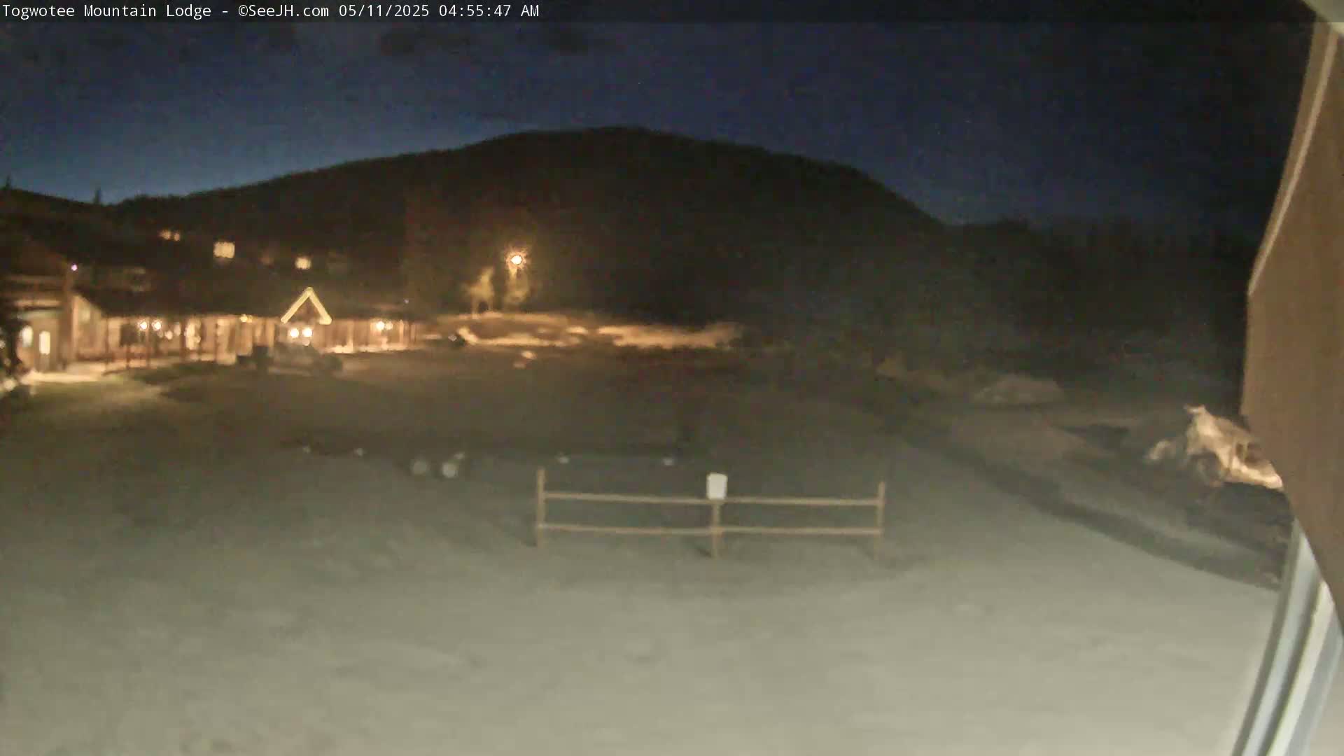 A nighttime view of a lodge illuminated against a dark mountain under a clear sky shows a snow-covered ground in front of it.